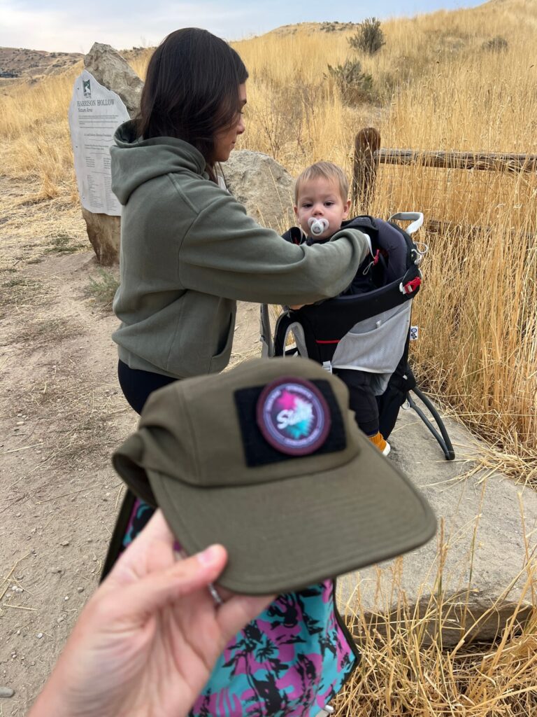 mom loading son into hiking backpack carrier with hat in forefront of photo