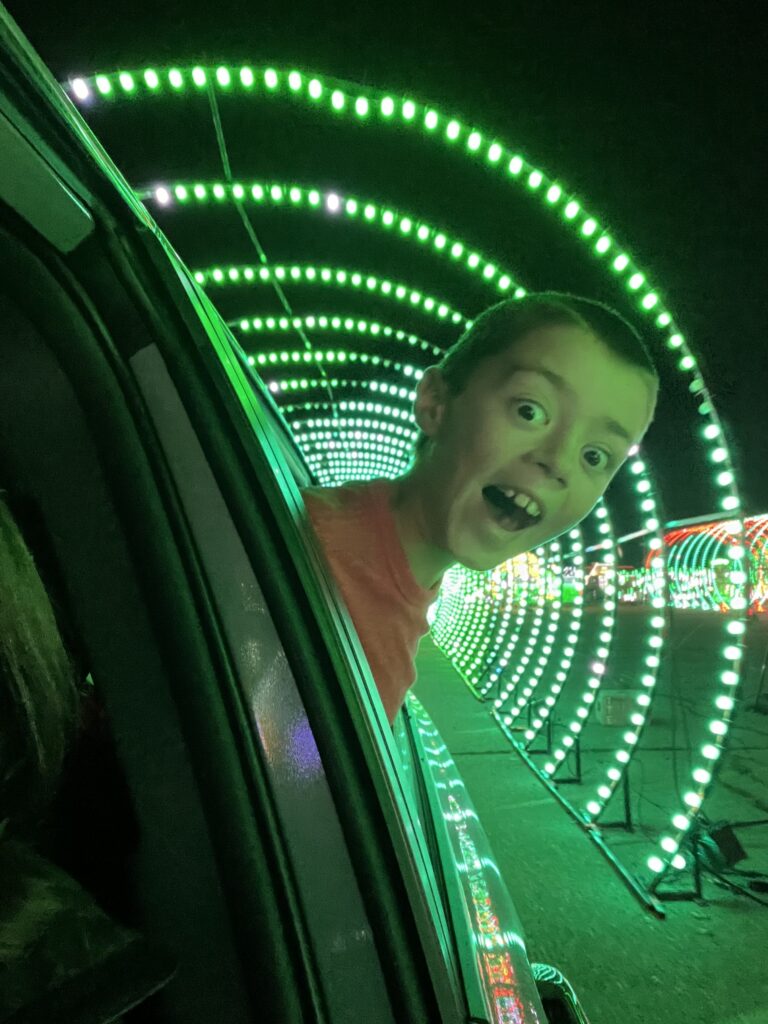 boy sticking head out car window with christmas lights behind