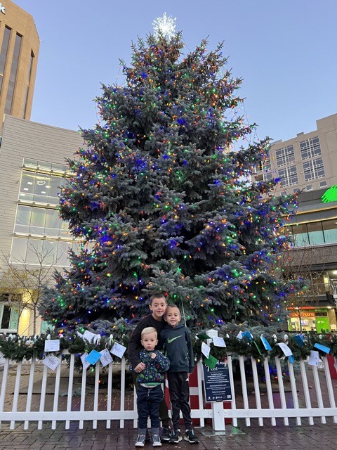 three boys standing in front of white fence around a giant christmas tree